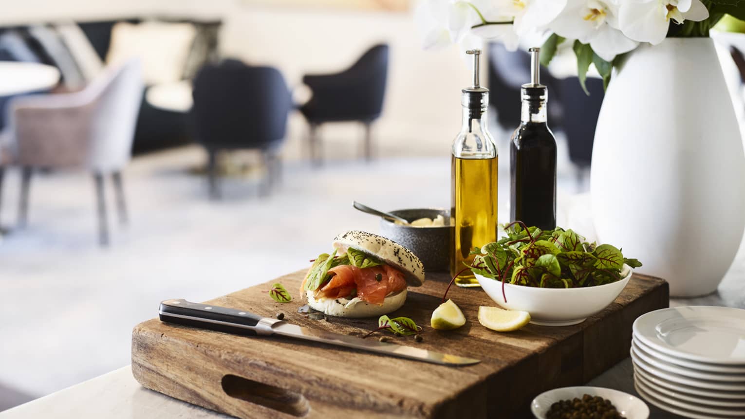 A bagel with lox, a bowl of greens, oil and vinegar bottles, and a knife on a wood cutting board by a vase of white flowers.