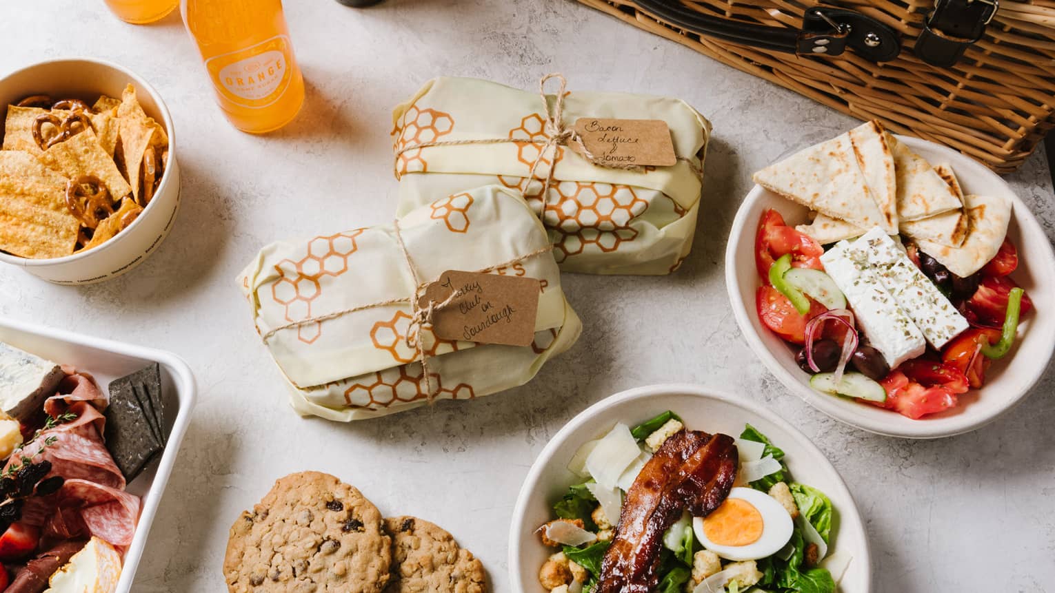An assortment of picnic food including salad, cookies, sandwiches, fruit, and sliced meat next to a brown wood basket.