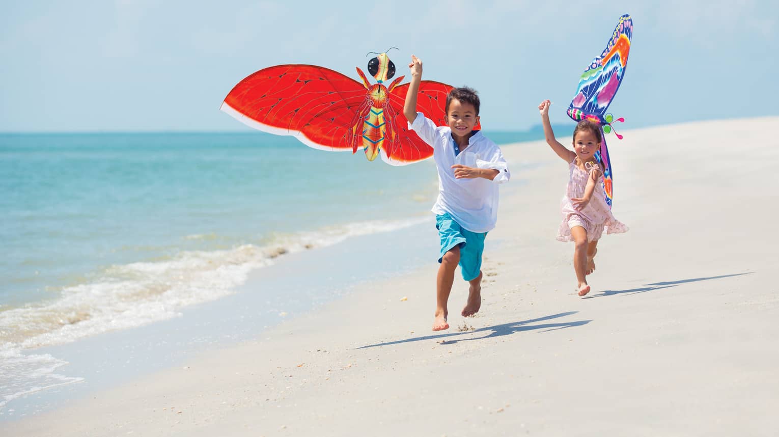 Two young children run with colourful butterfly kites on white sand beach