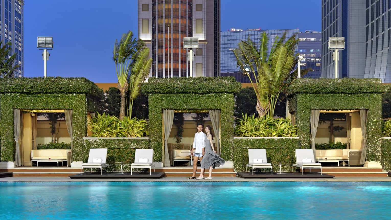 A couple smiles next to a pool, behind them a row of square archway cabanas covered in greenery and flanked by tall palms.