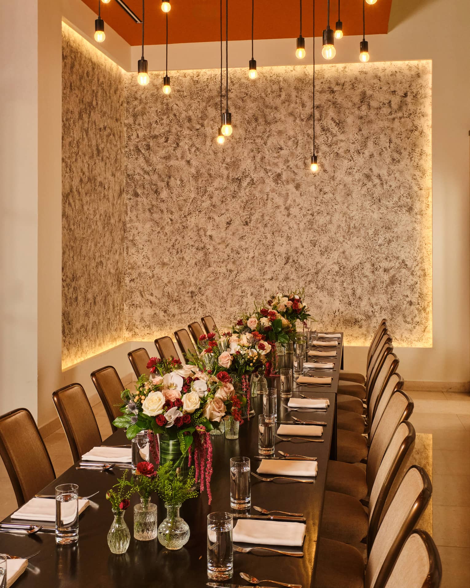 Long rectangular dining table set in a private dining room with red-and-light-pink floral arrangements in the centre and string lights overhead