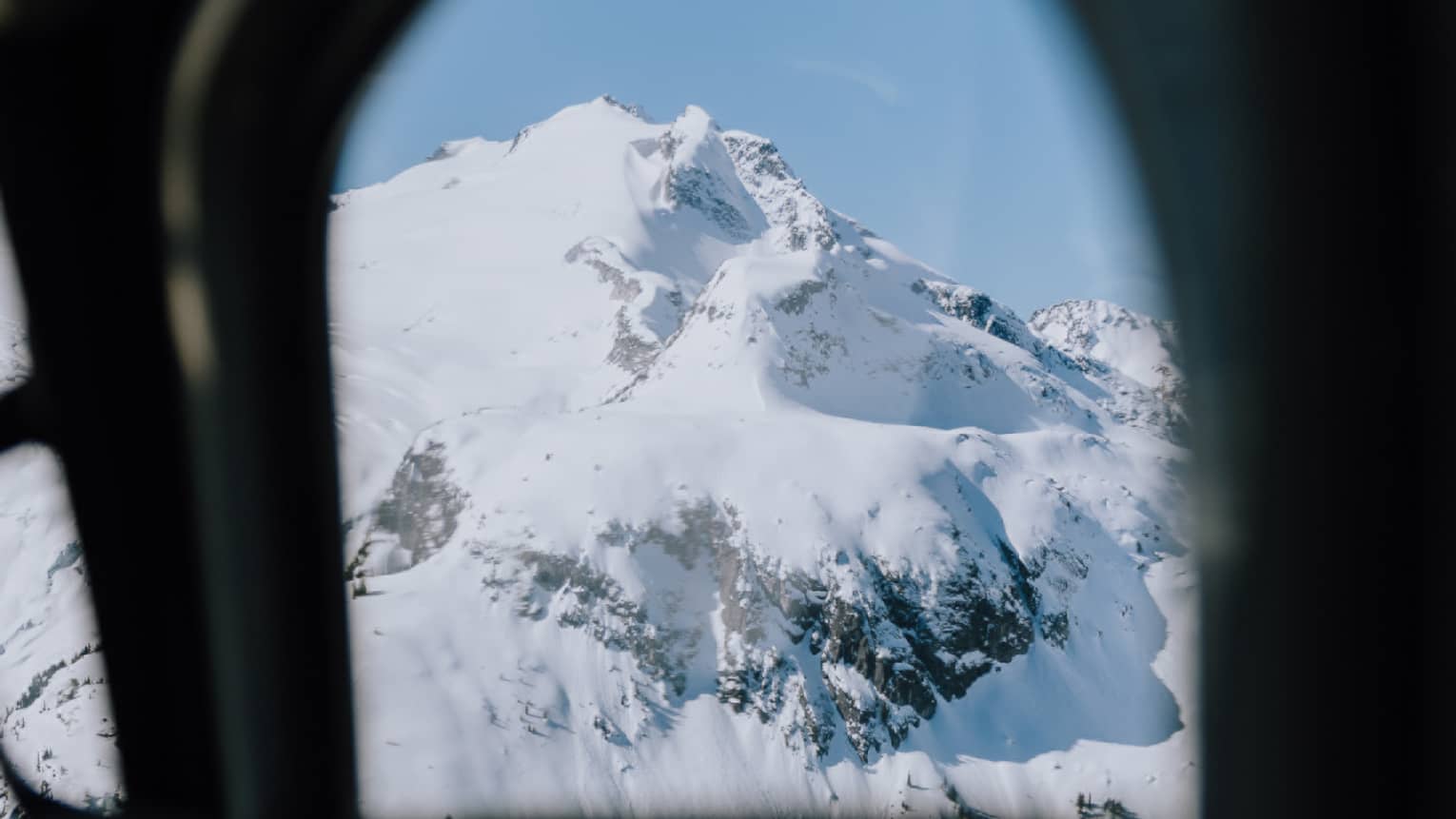 An aerial view through an airplane window of a mountain covered in snow with sporadic clumps of trees around the base.