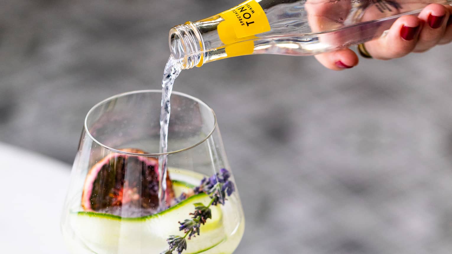 Hand pours bottle of sparkling water in wine glass with dried fruit, herbs