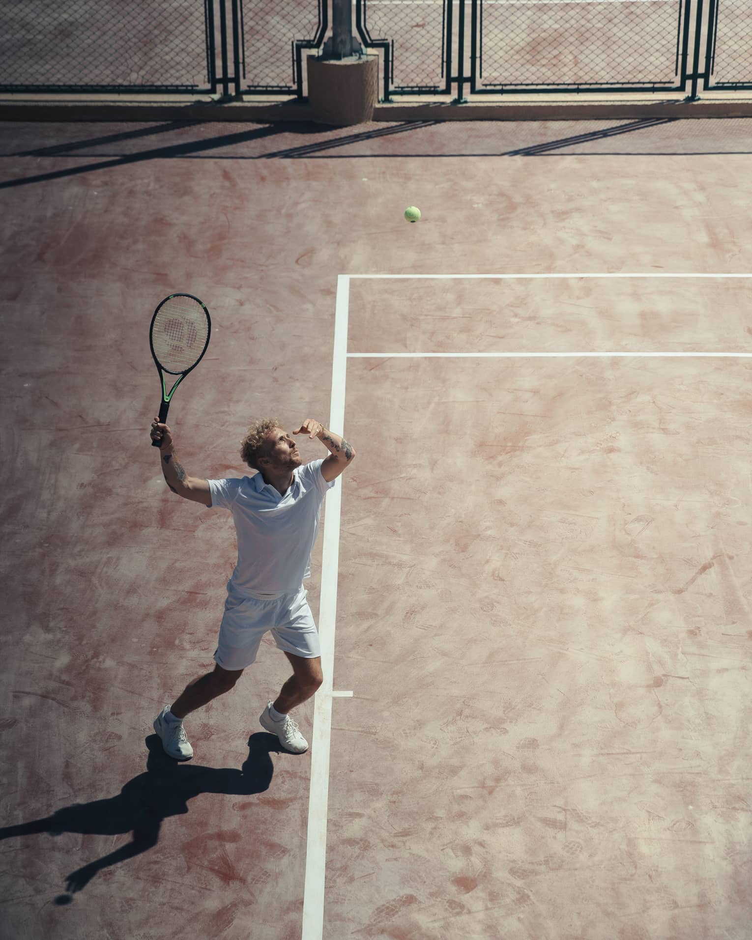 Man in white outfit playing tennis on reddish clay court
