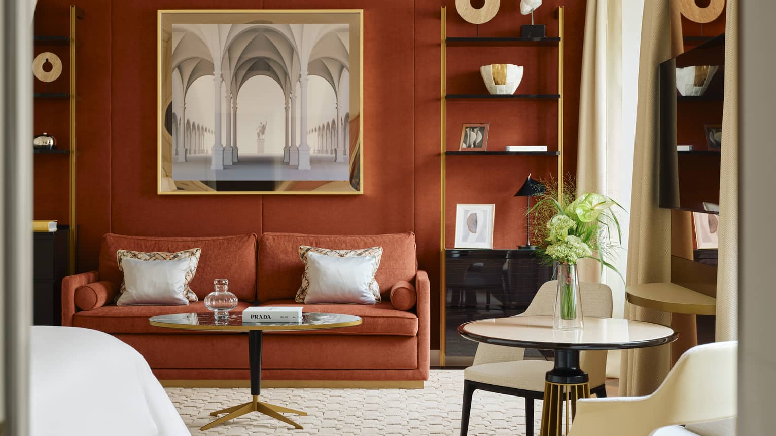 Contemporary hotel room with a dark burnt-orange accent wall and matching coloured sofa, two shelves containing artwork and crown molding