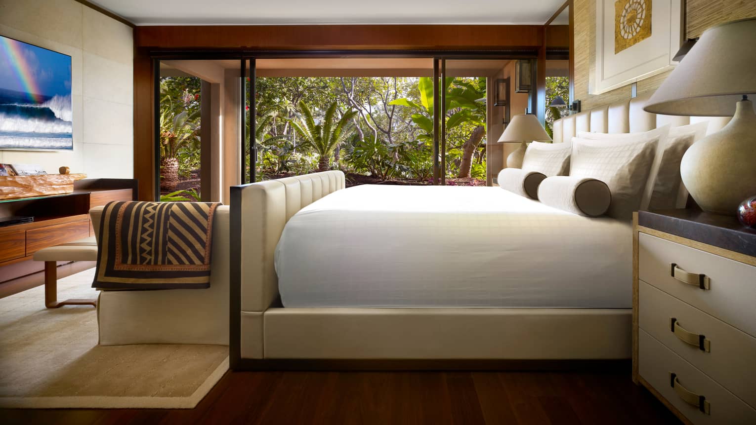 Four Seasons Garden-View Suite side of bed with white padded frame, throw blanket over small sofa, TV