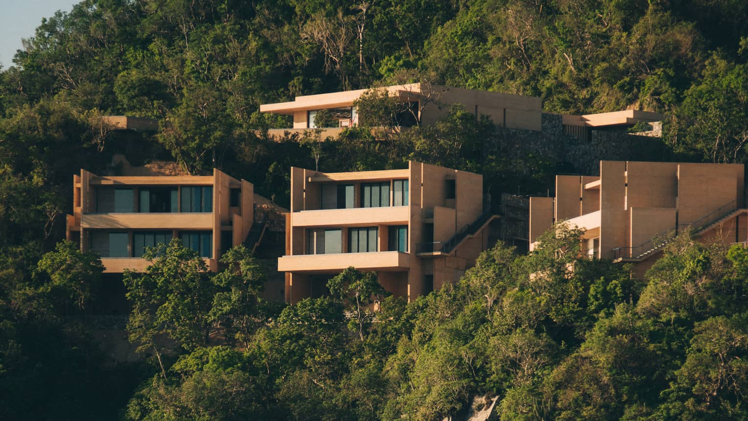 Modern cliffside villas surrounded by lush greenery, overlooking a secluded beach and ocean. The homes feature clean, angular designs, blending into the natural landscape.
