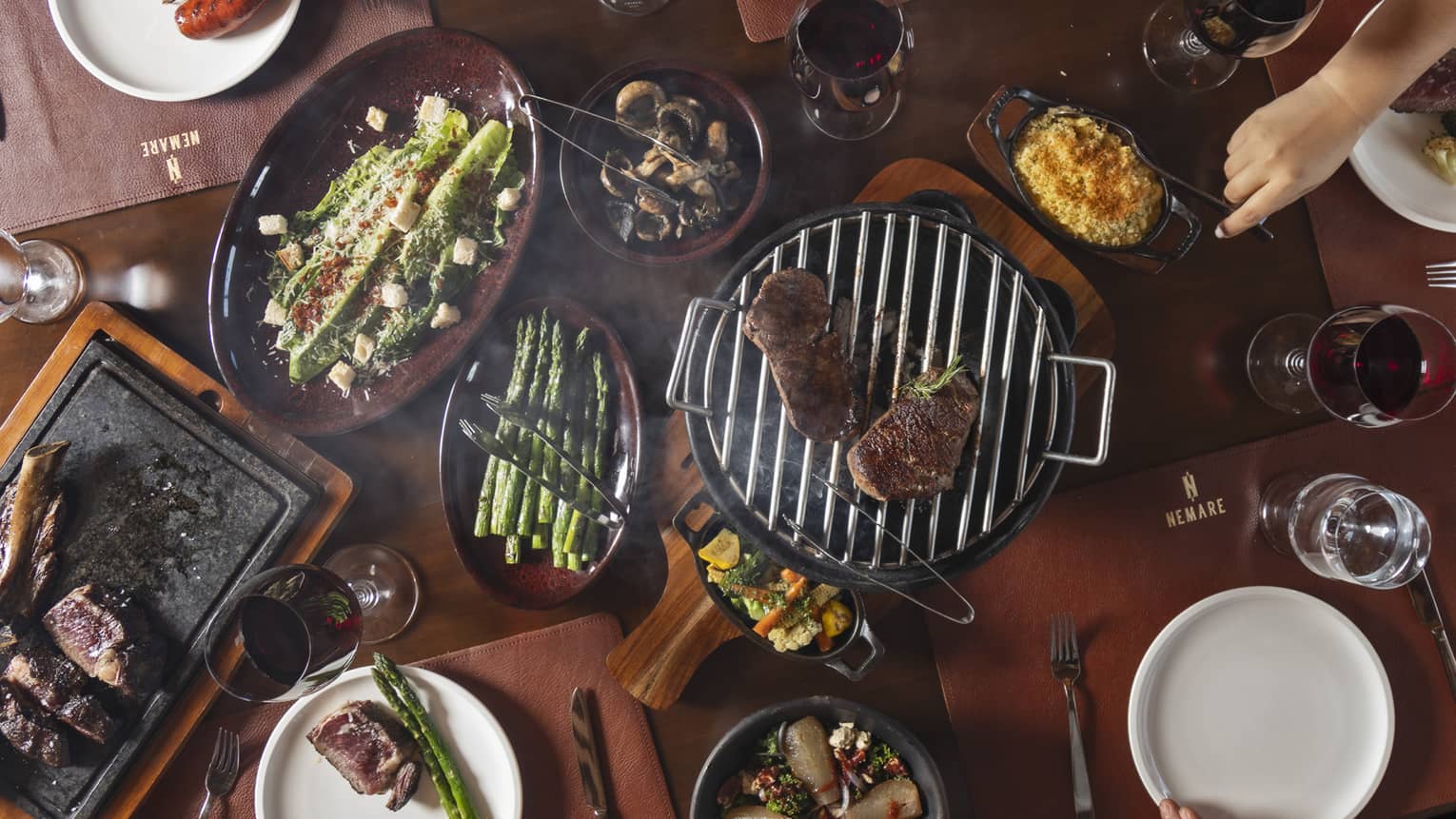 Overhead view of small grill placed in the centre of a dining table surrounded by various plates of food including asparagus, mushrooms, steak and more