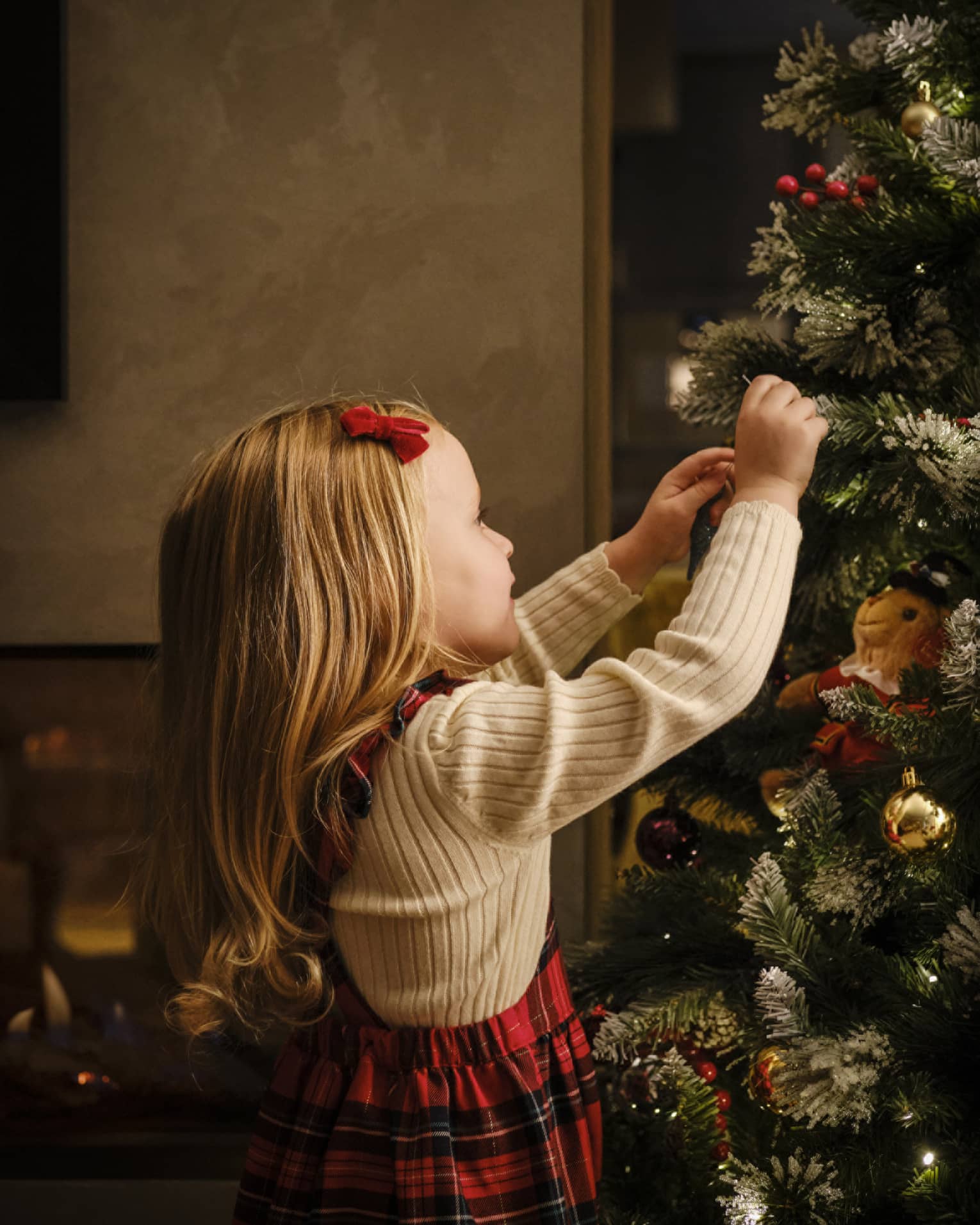 Close-up of a young child, red velvet barrette in their smooth blonde hair, arms reaching up to decorate a Christmas tree.