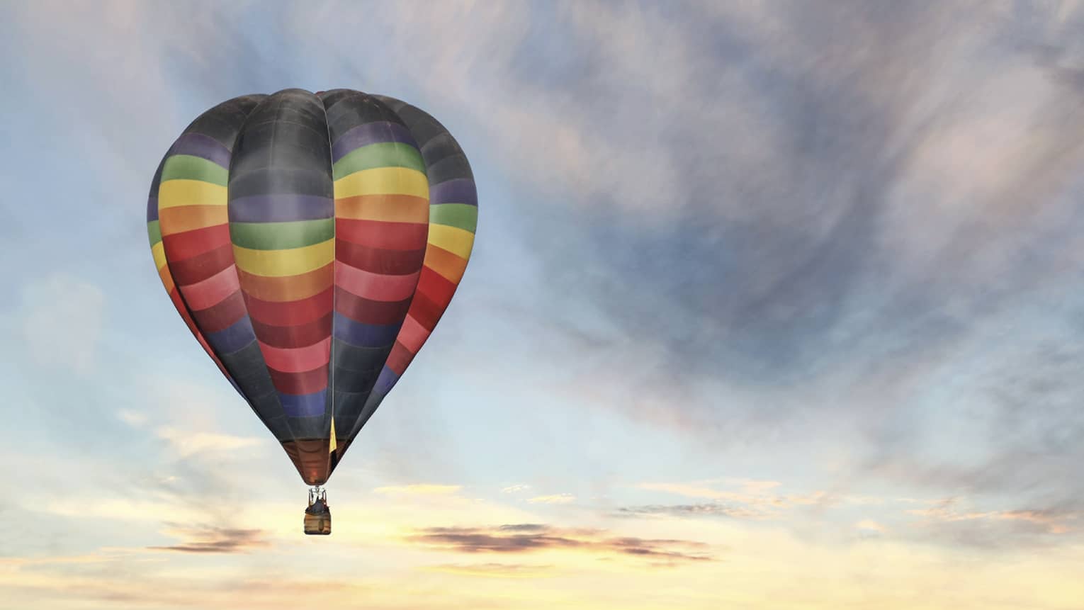 Rainbow-striped hot air balloon floats in the sky at sunset