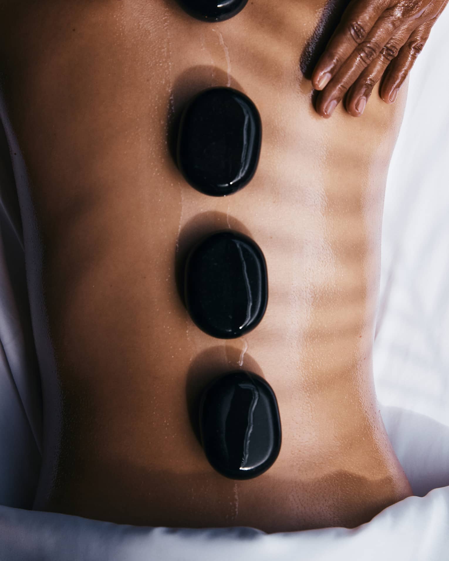 A person lying prone on a spa treatment table with smooth, round, black oval stones placed along their spine