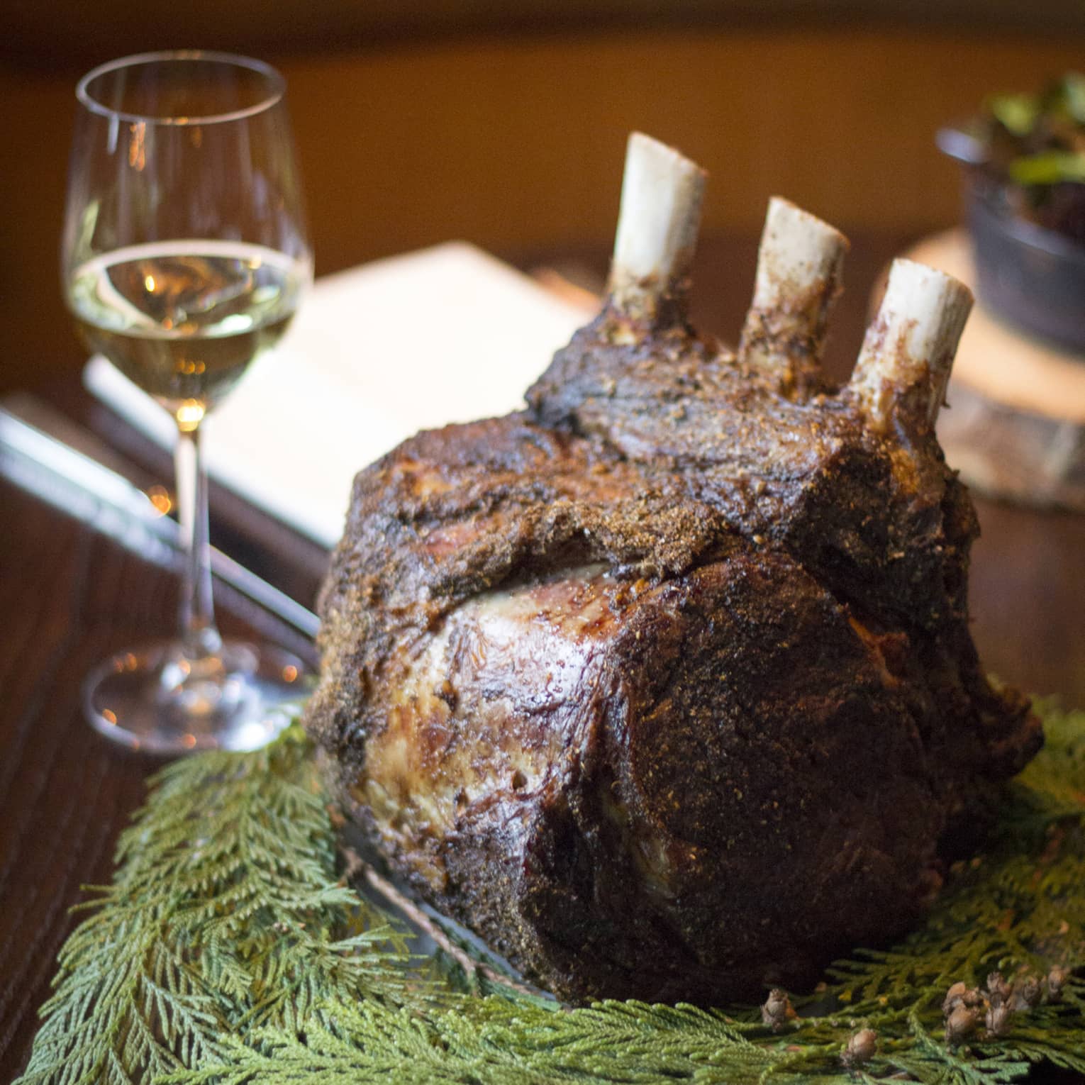A large bone in pork chop on herbs next to a glass of wine.