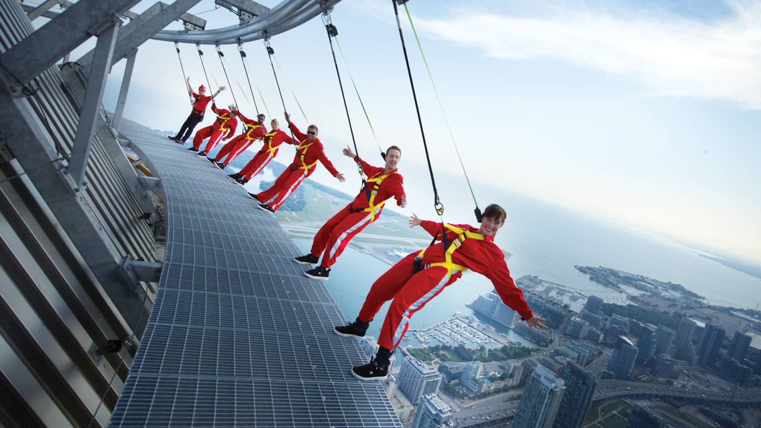 People wearing red jumpsuits, harnesses lean back over edge of CN Tower high above city