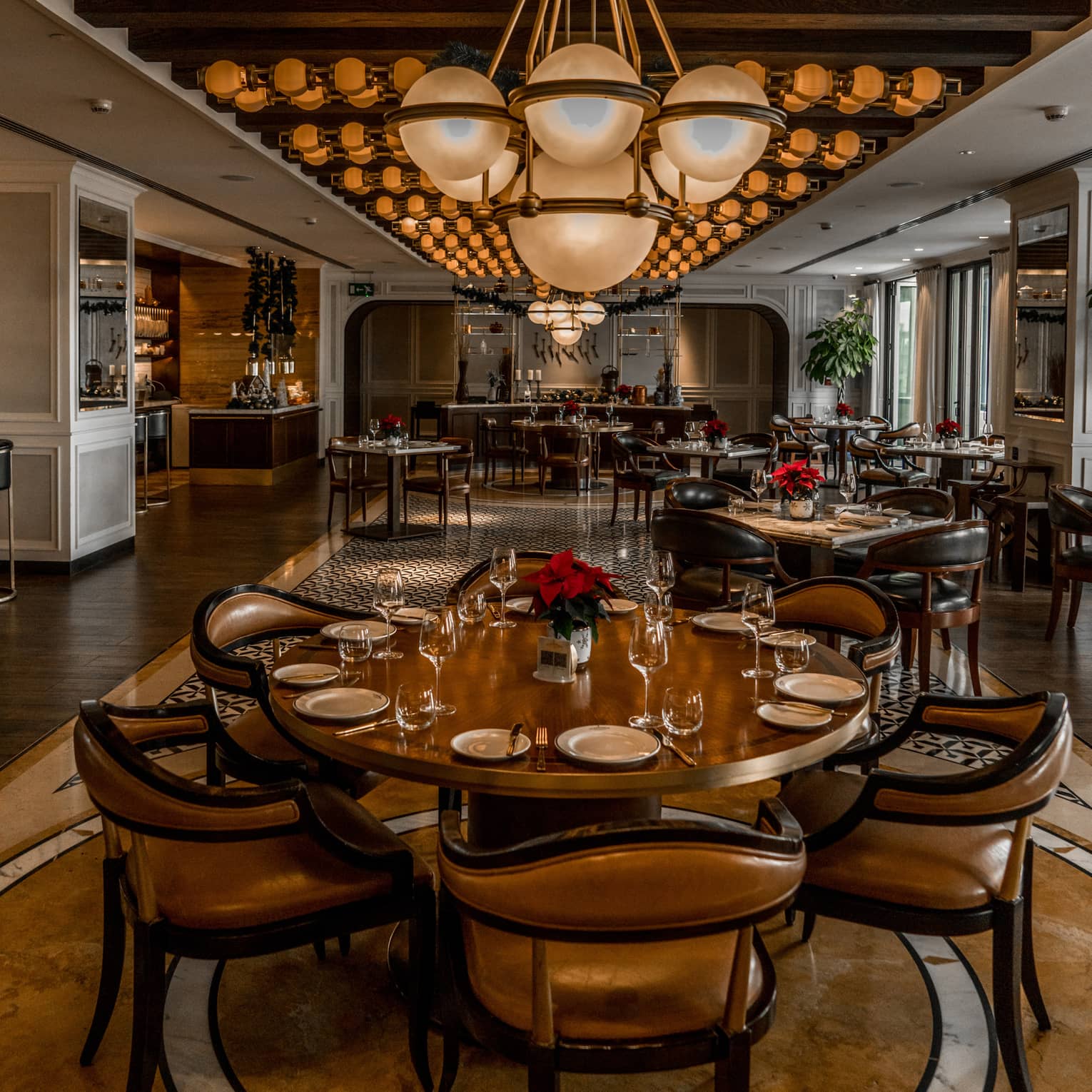 Brown leather dining chairs surround a round wooden table set for 6 in La Capital, with a globe-style light fixture hanging above