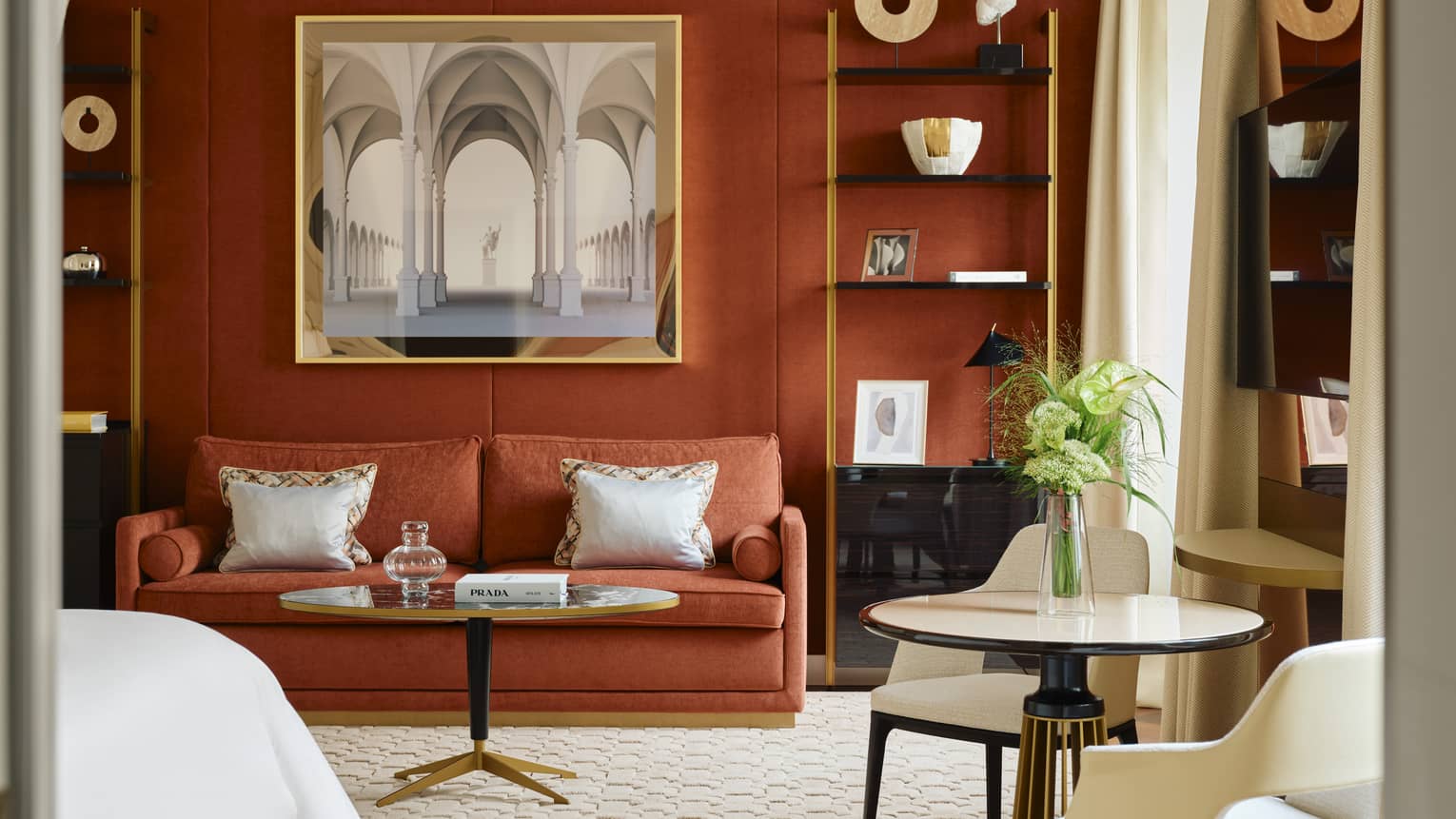 Contemporary hotel room with a dark burnt-orange accent wall and matching coloured sofa, two shelves containing artwork and crown molding