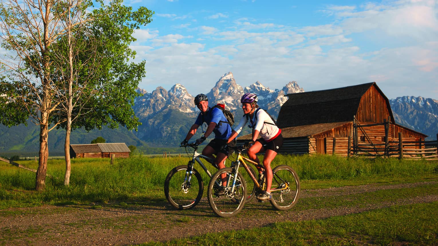 Man and woman on mountain bikes ride on path with log cabin and snow capped mountains in background