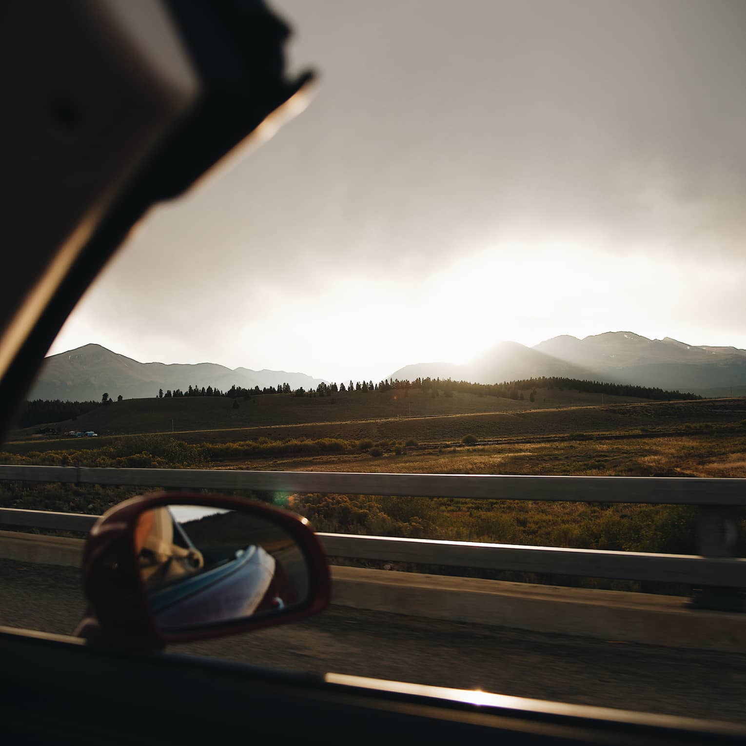 View from interior of passenger side mirror on a top down convertible, looking out to low distant mountains at sunset.