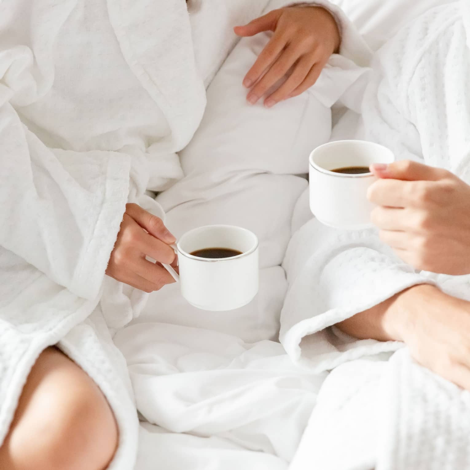 A faceless couple in white robes lounge on white bedding while holding mugs of black coffee.