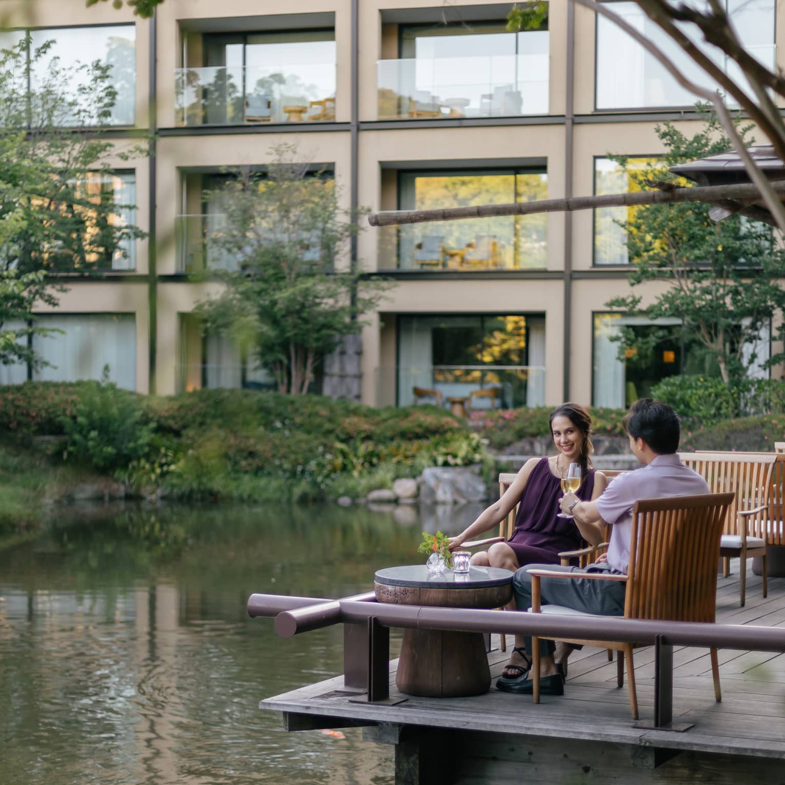 Smiling couple clink champagne flutes at a café table on a wood deck overlooking a rippling pond bordered by lush foliage.