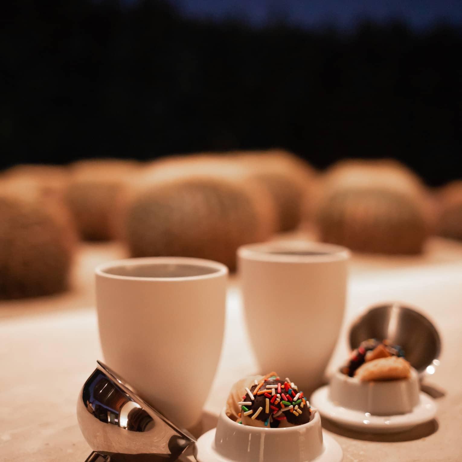Two coffee cups with mini stainless steel cloche domes beside bite sized desserts covered in chocolate and rainbow sprinkles.