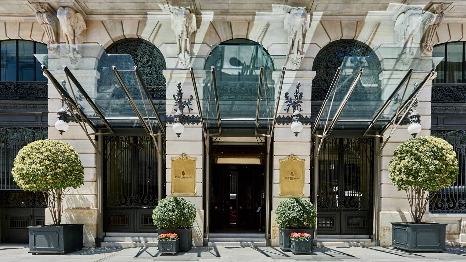 Exterior view of Hotel with potted foliage, glass canopy and golden plaques, second-story balcony visible