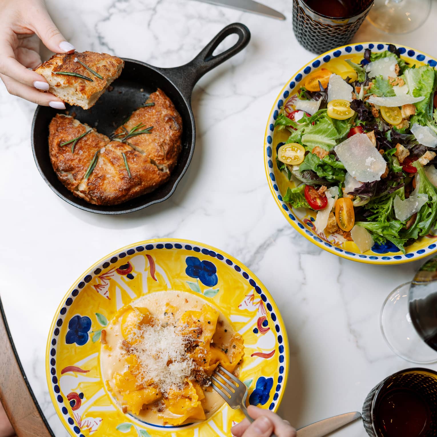 Amid colourful dishes, cutlery and glassware, a hand takes focaccia from an iron skillet, another takes a forkful of ravioli.
