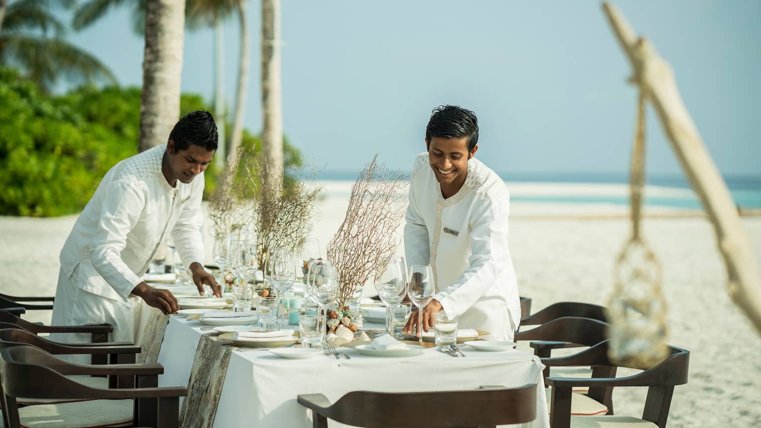 Two hotel staff set plates, wine glasses on private dining table on white sand beach