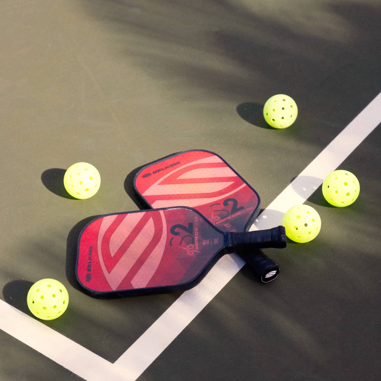 Close-up of two red and pink pickleball racquets, handles crisscrossed on a white court line, surrounded by five pickleballs.