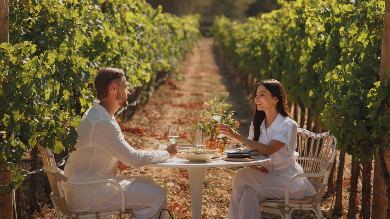 A smiling couple drink wine and dine at a small table set in a row of a sun-dappled and lush green vineyard.