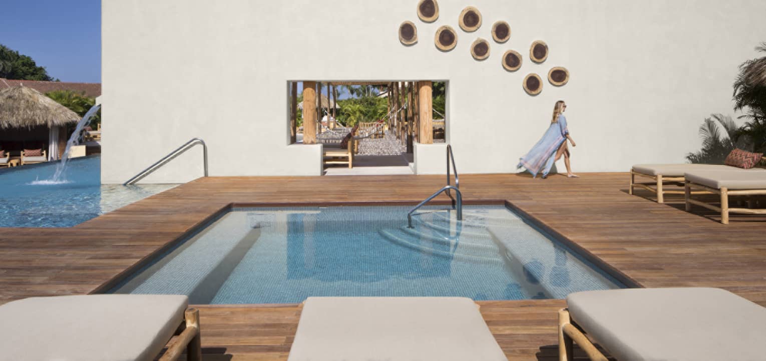 Woman wearing a long, flowy cover-up, walking past a stucco wall on a wooden pool deck