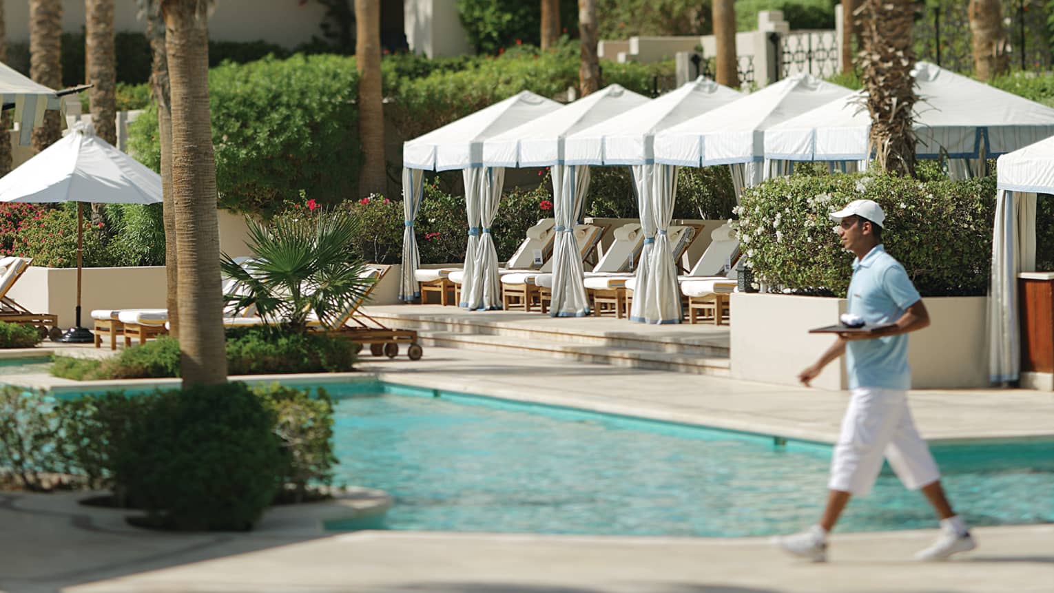 Server in uniform carries tray past outdoor swimming pool, white cabanas on sunny patio in background