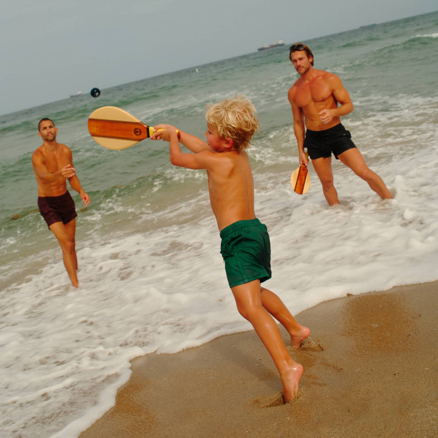 A young shild plays paddle ball in the surf with two adults