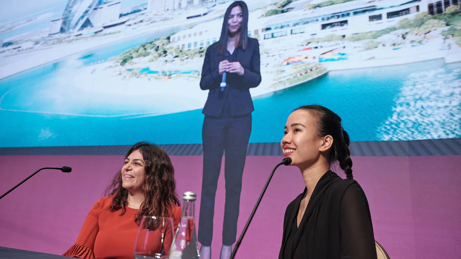 Presenters speaking at a table with large screen with additional presenter behind.
