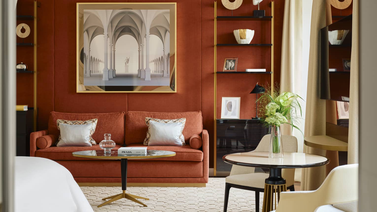 Contemporary hotel room with a dark burnt-orange accent wall and matching coloured sofa, two shelves containing artwork and crown molding