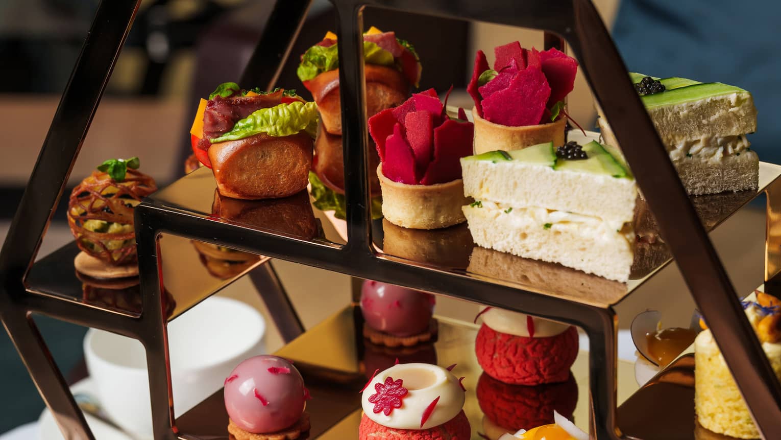 Bronze hexagonal three-tiered tray filled with small pastries and set on a table next to small containers of jam and a silver bread basket
