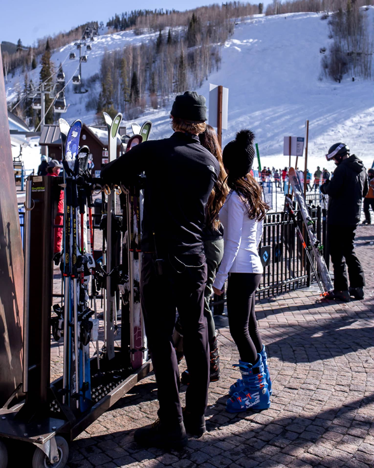 A group of people looking at skis and snowboards outside near a hill with snow on it.