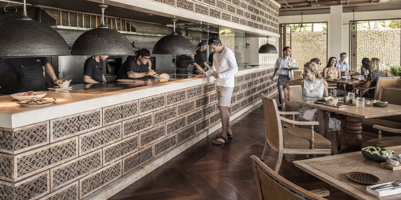 Restaurant guests watch as chefs plate entrees on ledge of open kitchen under accent wall