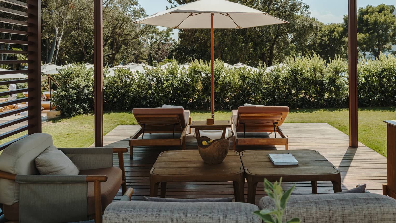 A shaded pool cabana with comfortable seating, a table, lounge chairs, an umbrella and lush green surroundings.
