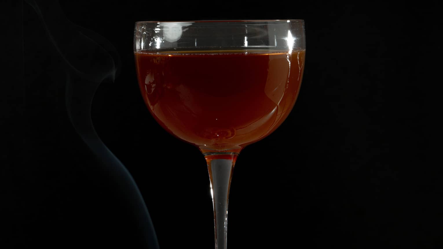 A dark amber cocktail in a stemmed glass with green garnishes around its base