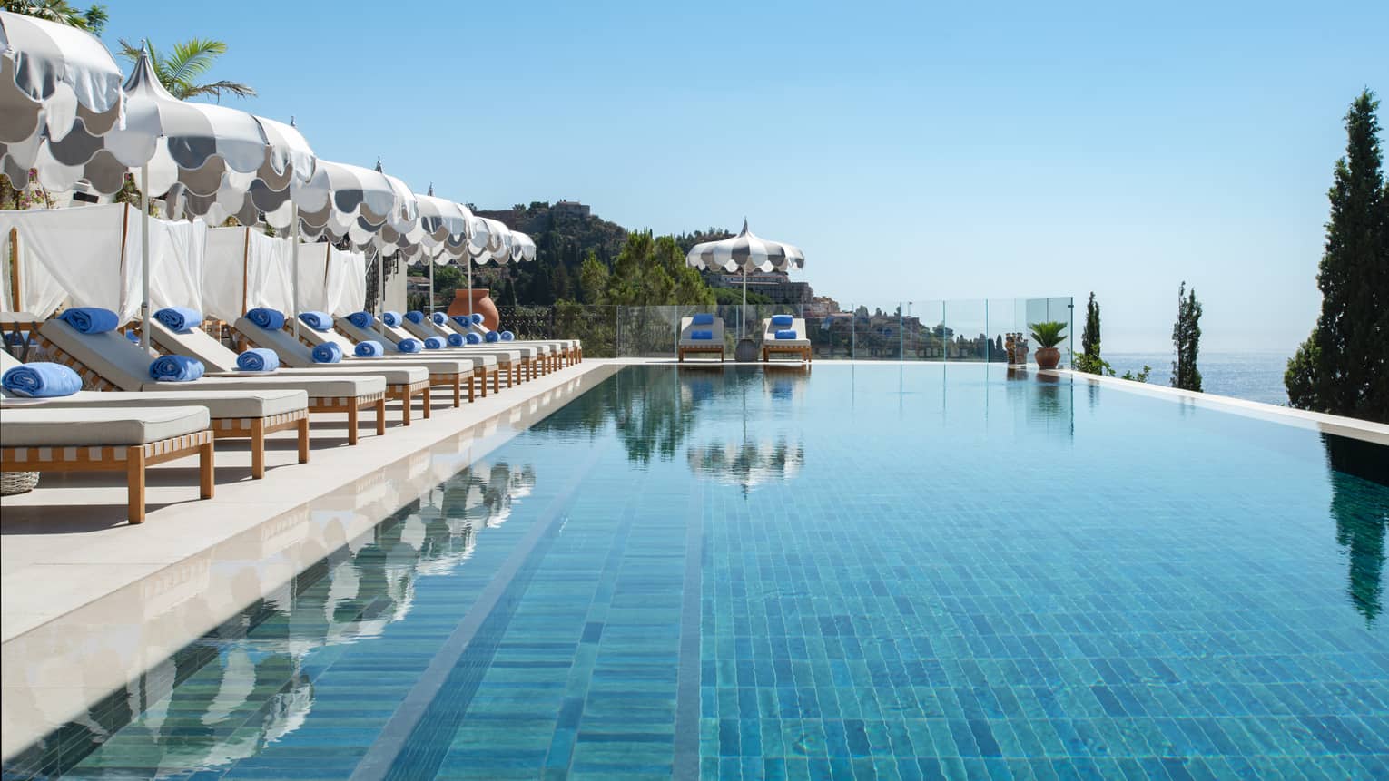A smooth inifinity-edge pool with lounge chairs and umbrellas along one side and end, and land and ocean views beyond it