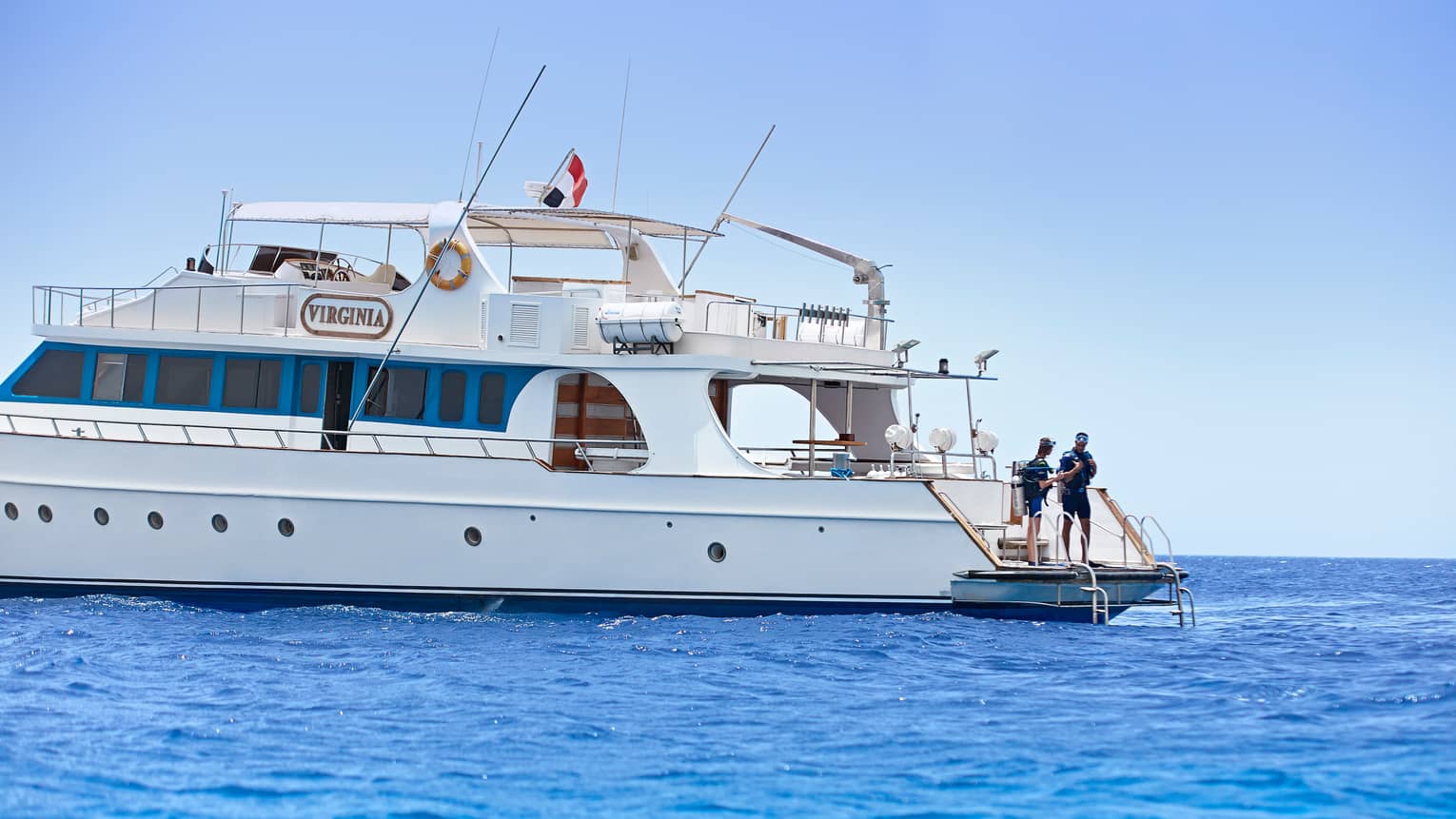 Two people wearing scuba gear prepare to jump into sea from back of large white yacht 