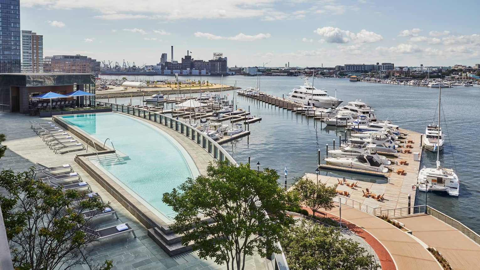A pool next to a dock with boats, a city can be sen in the distance across the water.