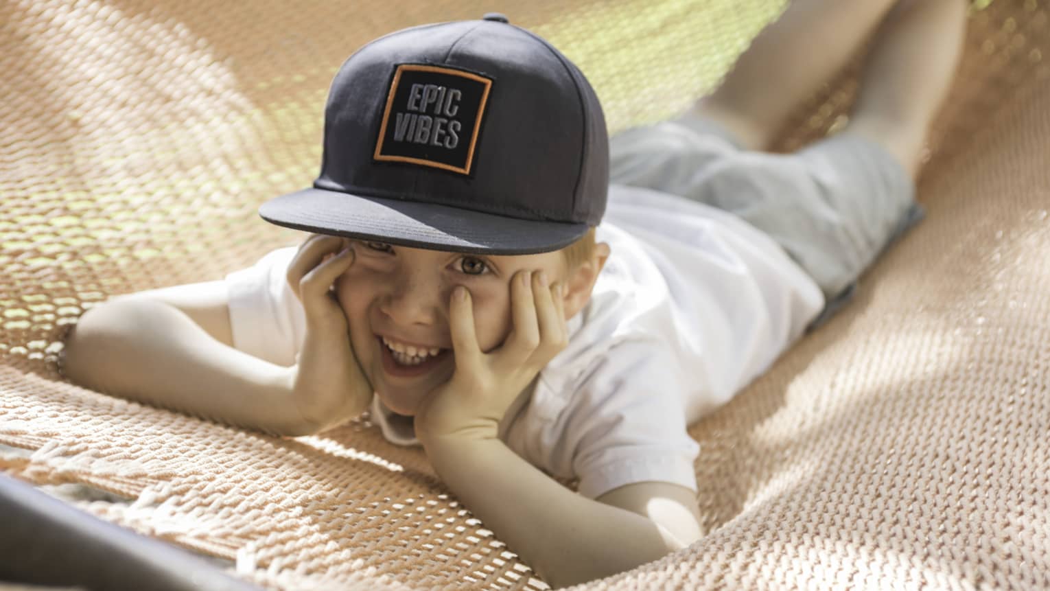 Child laying on hammock on stomach, face in hands and wearing a black cap