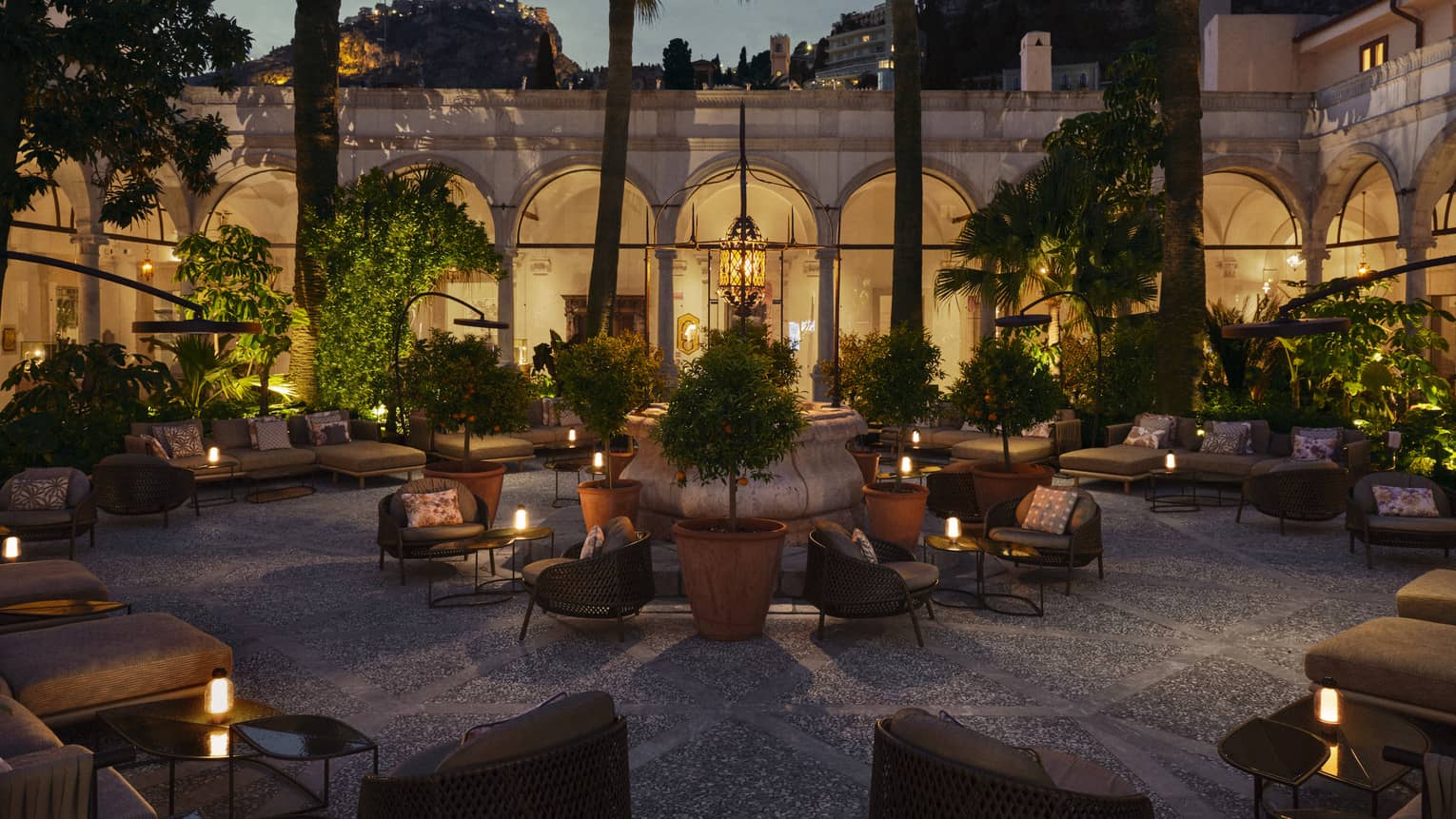 Courtyard-style lounge area with various seating areas and tables topped with candles, all set beneath some trees with an arched colonnade walkway in the background at night