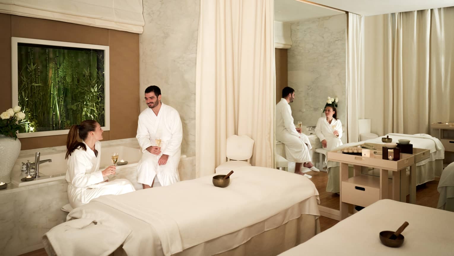 Robed couple beside tub and beds in couples spa treatment room with reflection in mirror