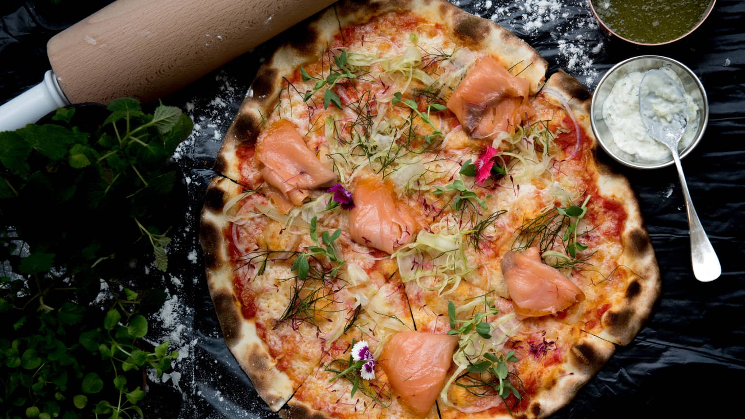 A wood-fired pizza topped with smoked salmon, fennel and dill, bowls of green and white sauces on the side. 