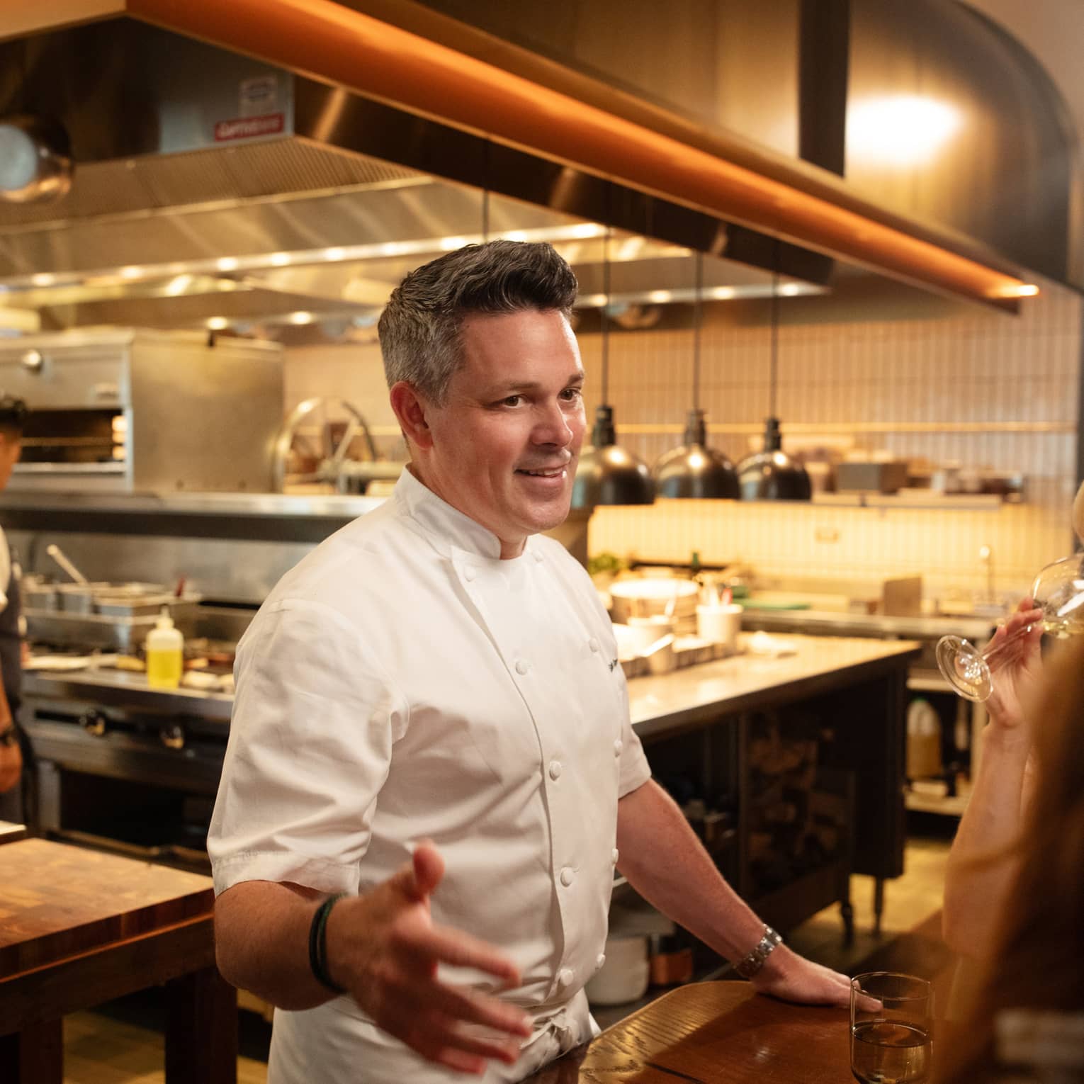 Chef standing beside a dining table in the kitchen talking to guests.