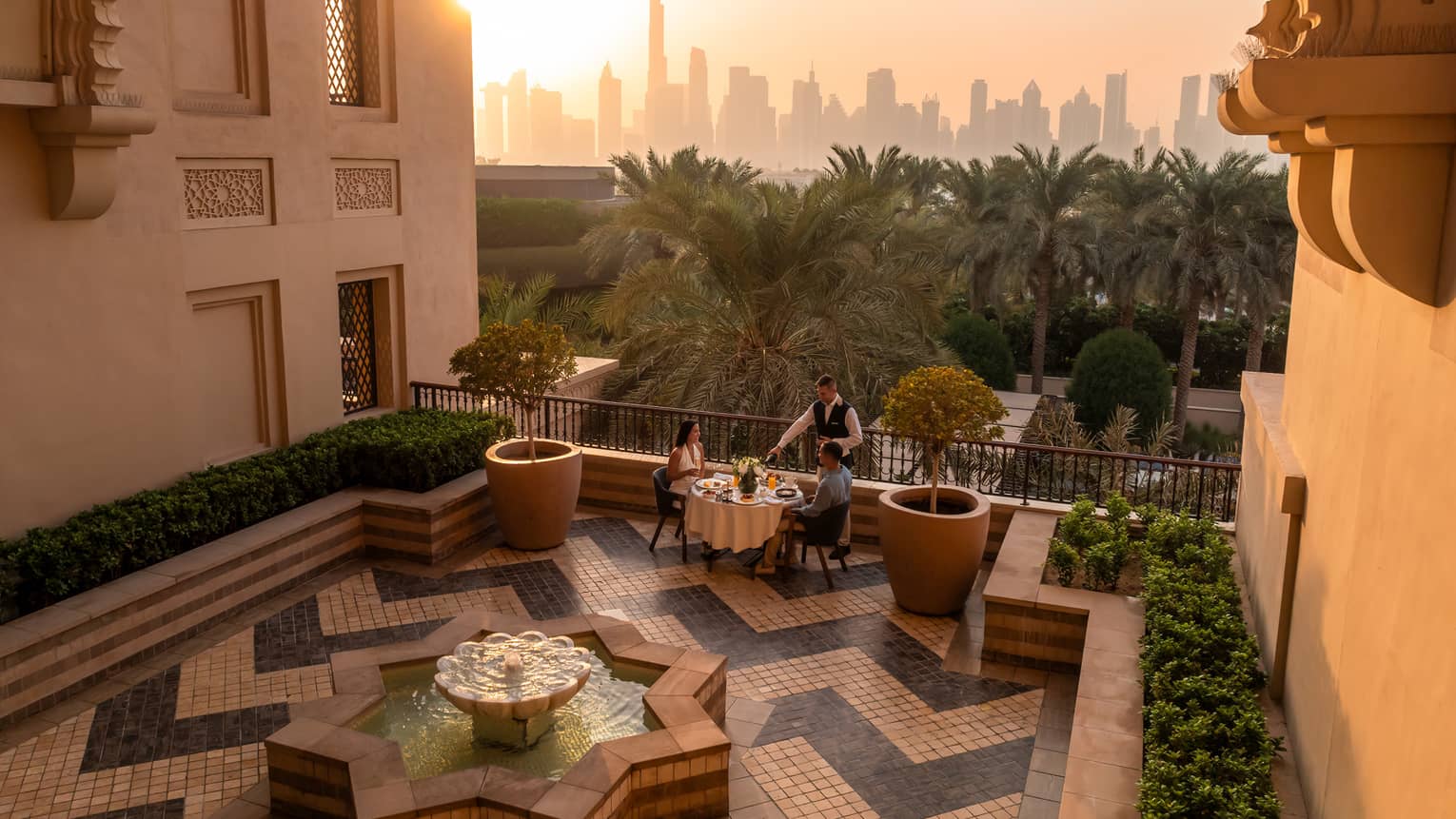 Table set for two on an open-air rooftop terrace with a chevron tiled floor and the sun rising over a city skyline in the near distance