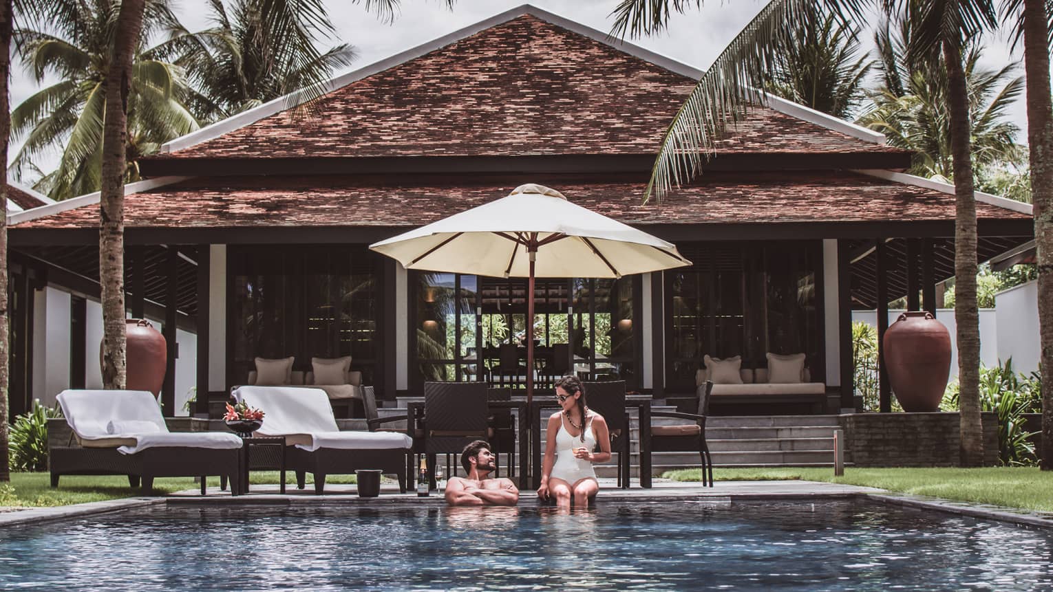 Man, woman in swimsuits at edge of outdoor pool under patio umbrella, by villa
