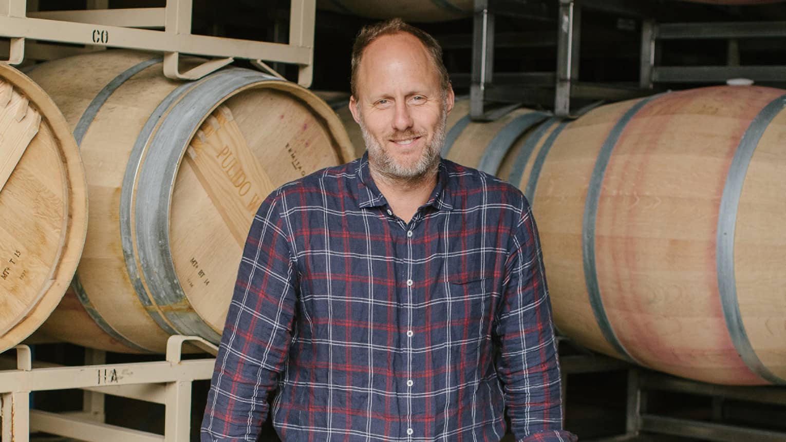 Thomas Brown stands before two racks of wine barrels as he rests his hands on two upright barrels and smiles at the camera.